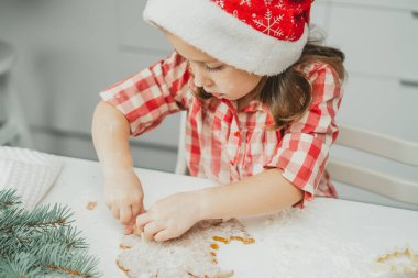 Kırmızı Noel şapkalı ve kareli gömlekli küçük siyah saçlı kız beyaz Noel süslemeli mutfakta sarılmış hamurdan zencefilli kurabiye kesiyor. Mutlu Noeller, mutlu yıllar.