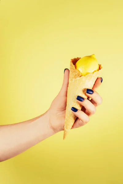 Female hand with blue nails holding waffle ice cream cone filled with lemon isolated on yellow. Vertical. Copy space