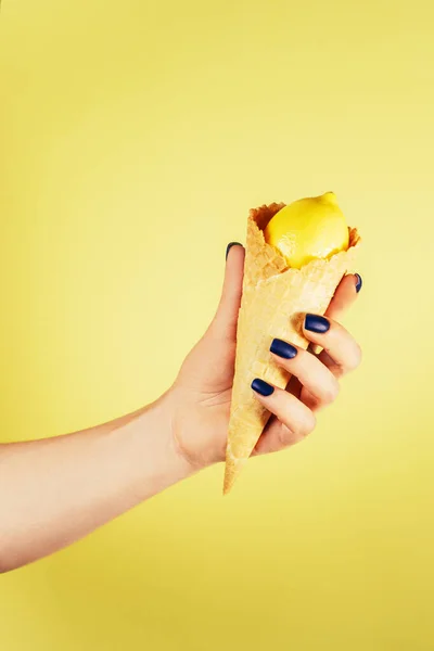 Mão feminina com unhas azuis segurando cone de sorvete waffle preenchido com limão isolado no amarelo. Vertical. Espaço de cópia — Fotografia de Stock
