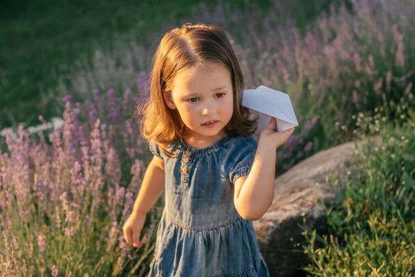 Little girl 3-4 with dark hair in denim dress in sun launches paper plane, among large bushes of lilac lavender