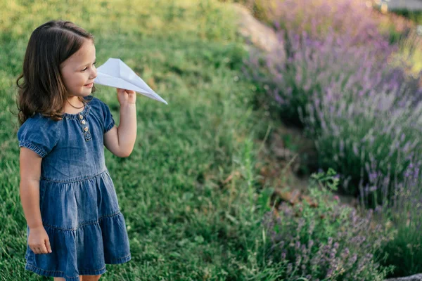 Little girl 3-4 with dark hair in denim dress in sun launches paper plane, among large bushes of lilac lavender