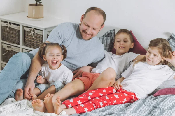 O pai brinca com três filhos na cama. O pai faz cócegas aos pés dos miúdos. Família de papai, duas meninas e menino — Fotografia de Stock