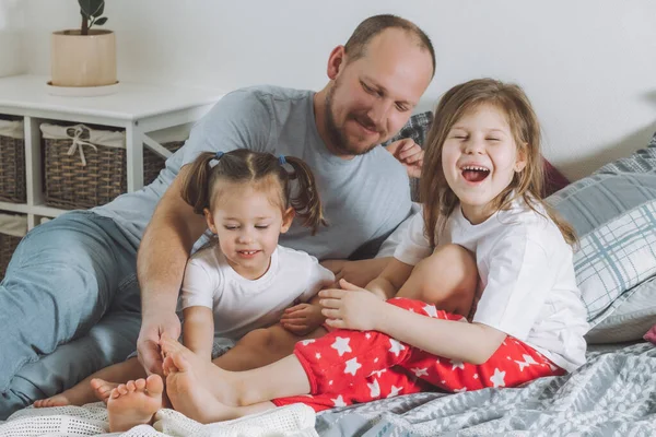 O pai brinca com dois filhos na cama. O pai faz cócegas aos pés dos miúdos. Família do pai, e duas meninas — Fotografia de Stock