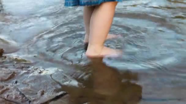 Legs of a little girl in a denim dress walking on the sand of a riverbank in the water in summer. Childhood. — Stockvideo