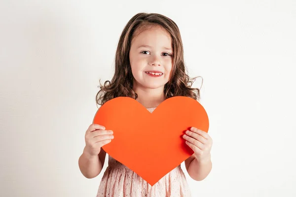 Portrait de petite fille aux longs cheveux bouclés tenant dans les mains grand coeur de papier rouge souriant sur fond blanc. Espace de copie. — Photo