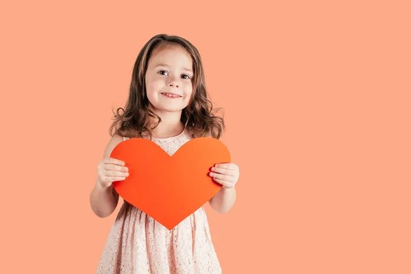 Retrato de menina com cabelo encaracolado longo segurar em mãos grande coração de papel vermelho sorrindo no fundo de pêssego. Espaço de cópia. — Fotografia de Stock