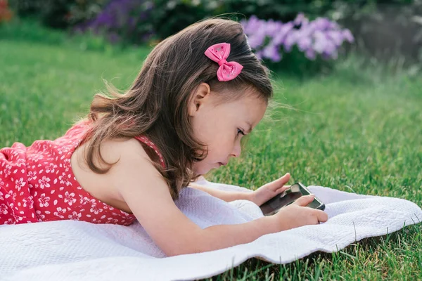 Portret van klein meisje 3-4 in het rood liggend op deken op groen gras en kijkend naar mobiele telefoon. Kinderen die gadgets gebruiken — Stockfoto