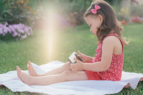 Klein 3-4 meisje in rode kleren zit op deken op groen gras en kijkt in mobiele telefoon. Kinderen met behulp van gadgets — Stockfoto