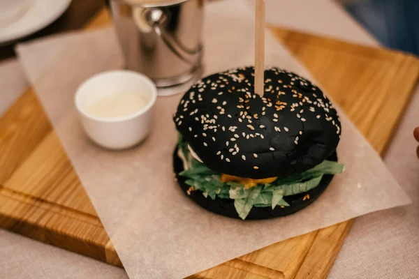 Sabroso delicioso sésamo negro hamburguesa de jamón con palo en ella, verde, lechuga, plato de salsa blanca. Café interior. De cerca. — Foto de Stock