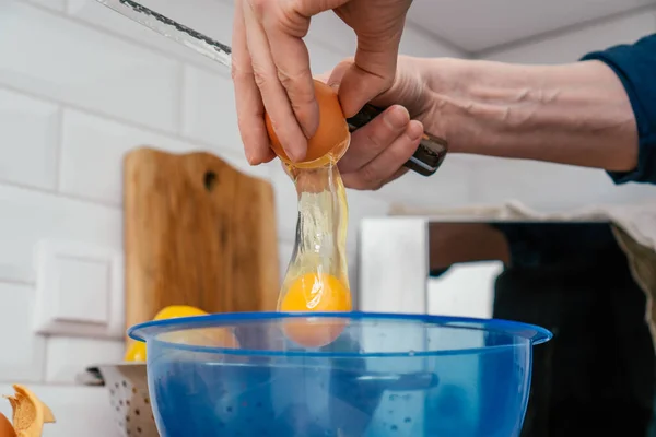 Крупним планом руки невпізнаваного чоловіка, який розбиває яйце ножем у блакитну пластикову миску поблизу сталевого друшляка. приготування їжі . — стокове фото
