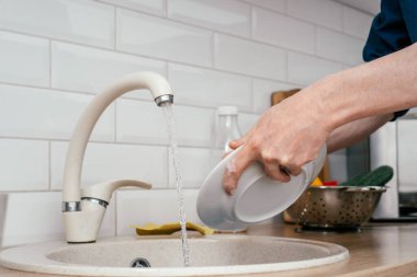 Mavi tişörtlü, tanınmayan bir adamı yakın çekimde bej mermer mutfak lavabosuyla akan suyun altında beyaz yuvarlak bir tabağa yıka..