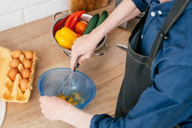 Mavi uzun kollu gömlekli, mavi plastik kasede yumurta çırpan, kırmızı, sarı dolma biberli çelik kevgir çırpan ve yumurtalı kutu açan tanınmamış bir adamın üst görüntüsü. Yemek pişirme.