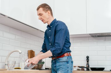 Orta yaşlı, kısa kızıl saçlı, mavi gömlekli bir adam bej mermer mutfak lavabosuyla beyaz mutfak lavabosunun altında beyaz modern mutfakta duran musluğun altında beyaz bir tabağı yıkıyor. Ev işi, temizlik konsepti.