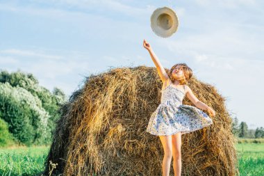 Mavi elbiseli küçük bir kızın portresi şapka takıyor, samanlığın yanında zıplıyor. Açık güneşli bulutlu bir gün. Neşe ve mutluluk konsepti. Arka planda mavi açık gökyüzü. Rüzgar hasır şapkayı alıp götürüyor. Düşük açı