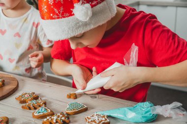 8 yaşındaki kırmızı Noel Baba şapkalı bir çocuk Noel kurabiyelerini mutfak masasında ahşap kremayla süslüyor. Mutlu Noeller, mutlu yıllar.