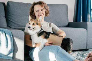Kıvırcık saçlı, neşeli, çıplak ayaklı, köpek Corgi 'si olan, rahat oturma odasındaki halıda oturup kitap okuyan bir kadın. Sakin ve rahat bir atmosfer. Hafta sonu tatili