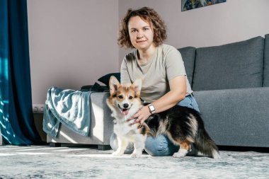 Kıvırcık saçlı, mutlu, çıplak ayaklı, köpek Corgi 'si olan, rahat oturma odasında, kanepenin yanında oturan, dinlenen ve oynayan bir kadın. Aktif ve sağlıklı bir hayvan, sağlık hizmeti. Hafta sonu boş zaman aktivitesi