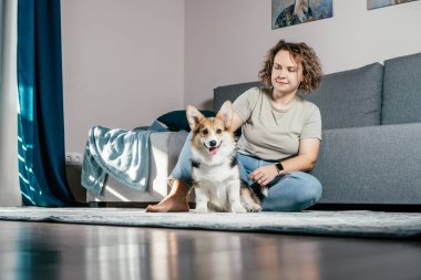 Kıvırcık saçlı, mutlu, çıplak ayaklı, köpek Corgi 'si olan, rahat oturma odasında, kanepenin yanında oturan, dinlenen ve oynayan bir kadın. Aktif ve sağlıklı bir hayvan, sağlık hizmeti. Hafta sonu boş zaman aktivitesi