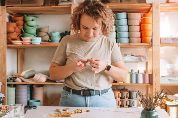 Doğal güvenli aksesuarlar yapan bir kadın. Masanın üzerinde, eko tarzı. Boncukları diziyorum. Ivır zıvır çıngırağı süslemeli bir oyuncak. Bebek dişleri çiğnemek için el yapımı oyuncaklar. Adım adım. Çocuk mağazası