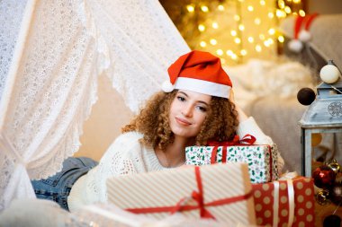 Kıvırcık saçlı, kırmızı Noel şapkalı, elinde hediye kutusu olan genç bir kadın.