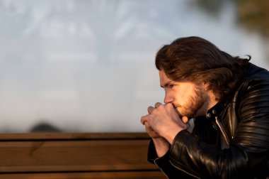 Ciddi ciddi sakallı adam siyah deri ceket giyiyor ve dışarıda oturup düşünüyor.