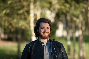 Mutlu genç sakallı adam gülümsüyor ve sonbahar ağaçlarının arka planında kameraya bakıyor.