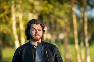 Genç sakallı adam dikilip kameraya bakıyor. Renkli sonbahar ağaçlarının arka planında.