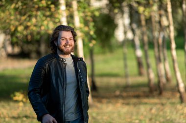 Mutlu genç sakallı adam gülümsüyor ve sonbahar ağaçlarının arka planında kameraya bakıyor.