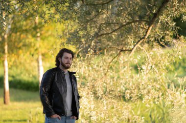 Sonbahar parkında ağaçların yanında duran sakallı genç bir adam düşünceli bir şekilde uzaklara bakıyor.