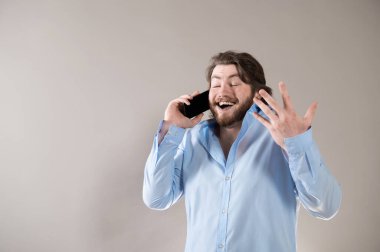 Genç sakallı adam, gri arka planda izole edilmiş bir şekilde telefonla arıyor ve zevkle konuşuyor.