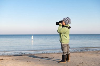 Elinde fotoğraf makinesi olan küçük çocuk sahilde dururken denizin fotoğraflarını çekiyor.