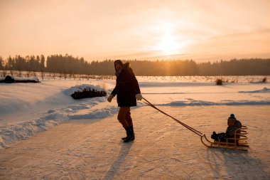 Baba, kışın açık havada kızakla gezerken çocuğu çekiyor. Baba oğluyla birlikte kızakla gölde yürüyor.