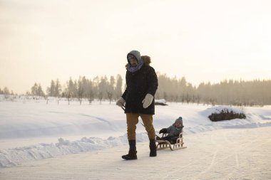 Baba, kışın açık havada kızakla gezerken çocuğu çekiyor. Güzel akşam güneşi.