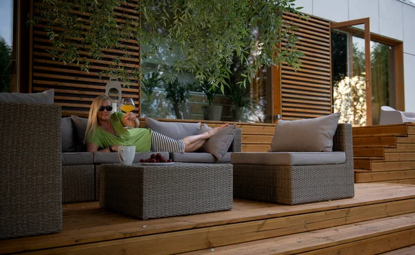 Hoş, orta yaşlı, mutlu bir kadın yaz terasında dinleniyor. Taze meyve suyu kokteyli içiyor ve olumlu duygular besliyor.