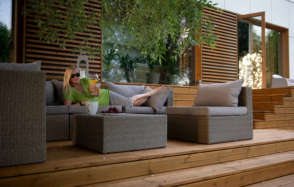 Hoş, orta yaşlı, mutlu bir kadın yaz terasında dinleniyor. Taze meyve suyu kokteyli içiyor ve olumlu duygular besliyor.