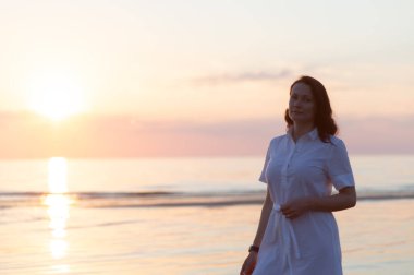 Güzel olgun bir kadın sahilde durup düşünceli bir şekilde kameraya bakar.
