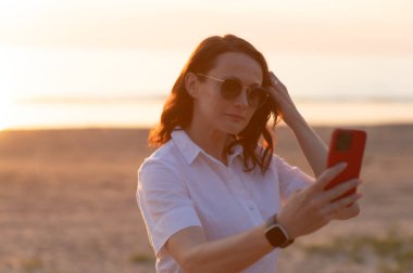 Gün batımında sahilde beklerken akıllı telefondan selfie çeken genç bir kadın.