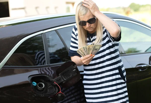 Desperate woman hold cash dollar bills next to car with an open tank, concept of rising fuel prices