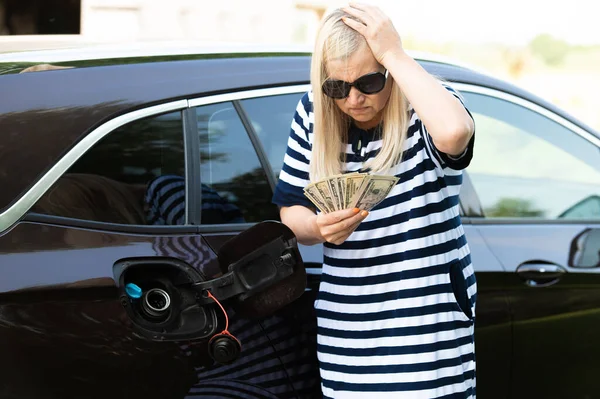 Desperate woman hold cash dollar bills next to car with an open tank, concept of rising fuel prices