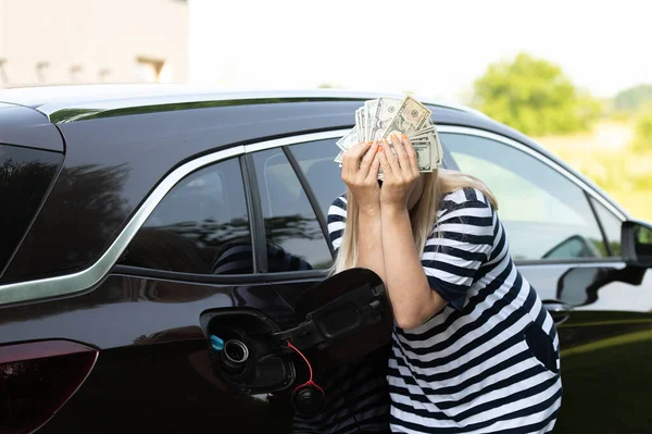 Desperate woman hides face behind cash near an open car fuel tank, concept of rising fuel prices