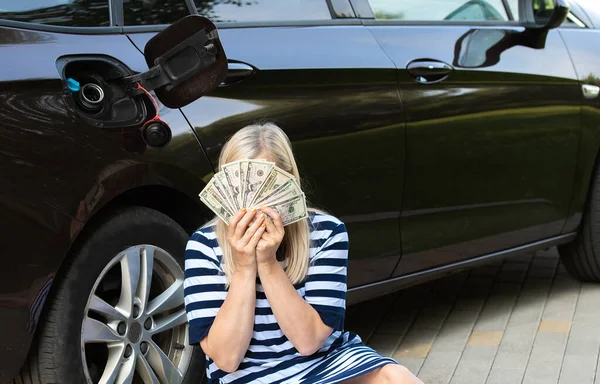 Desperate woman hides face behind cash near an open car fuel tank, concept of rising fuel prices