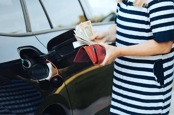 woman takes money dollars out of her wallet while standing next to a car with an open fuel tank