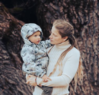 Annesinin kollarında bebekle doğadaki büyük ağacın önünde dikilirken görüntüsü.
