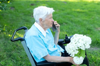 Yaşlı beyaz kadın bahçede tekerlekli sandalyede cep telefonu kullanıyor.