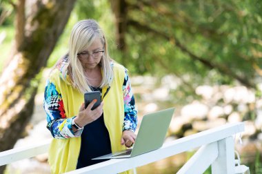İş parkında ayakta duran ve cep telefonuna mesaj yazarken bilgisayarını kullanan kadının portresi. Çevrimiçi çalışan iş kadını.