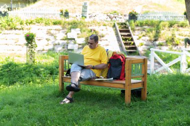 Orta yaşlı bir adam parktaki ahşap bankta oturuyor ve laptopunu kullanıyor, dışarıda çalışıyor.
