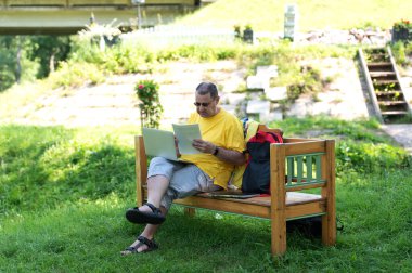 Orta yaşlı bir adam parktaki ahşap bankta oturuyor ve bilgisayarını kullanıyor, dışarıda belgelerle çalışıyor.