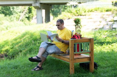 Orta yaşlı bir adam parktaki ahşap bankta oturuyor ve bilgisayarını kullanıyor, dışarıda belgelerle çalışıyor.
