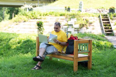 Orta yaşlı bir adam parktaki ahşap bankta oturuyor ve bilgisayarını kullanıyor, dışarıda belgelerle çalışıyor.