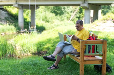 Orta yaşlı bir adam parktaki ahşap bankta oturuyor ve bilgisayarını kullanıyor, dışarıda belgelerle çalışıyor.